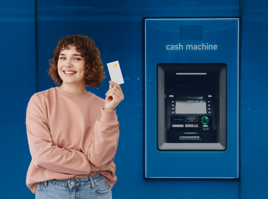 woman standing in a pink jumper and blue trousers happy holding a shite bank card in front of cash machine from a bank